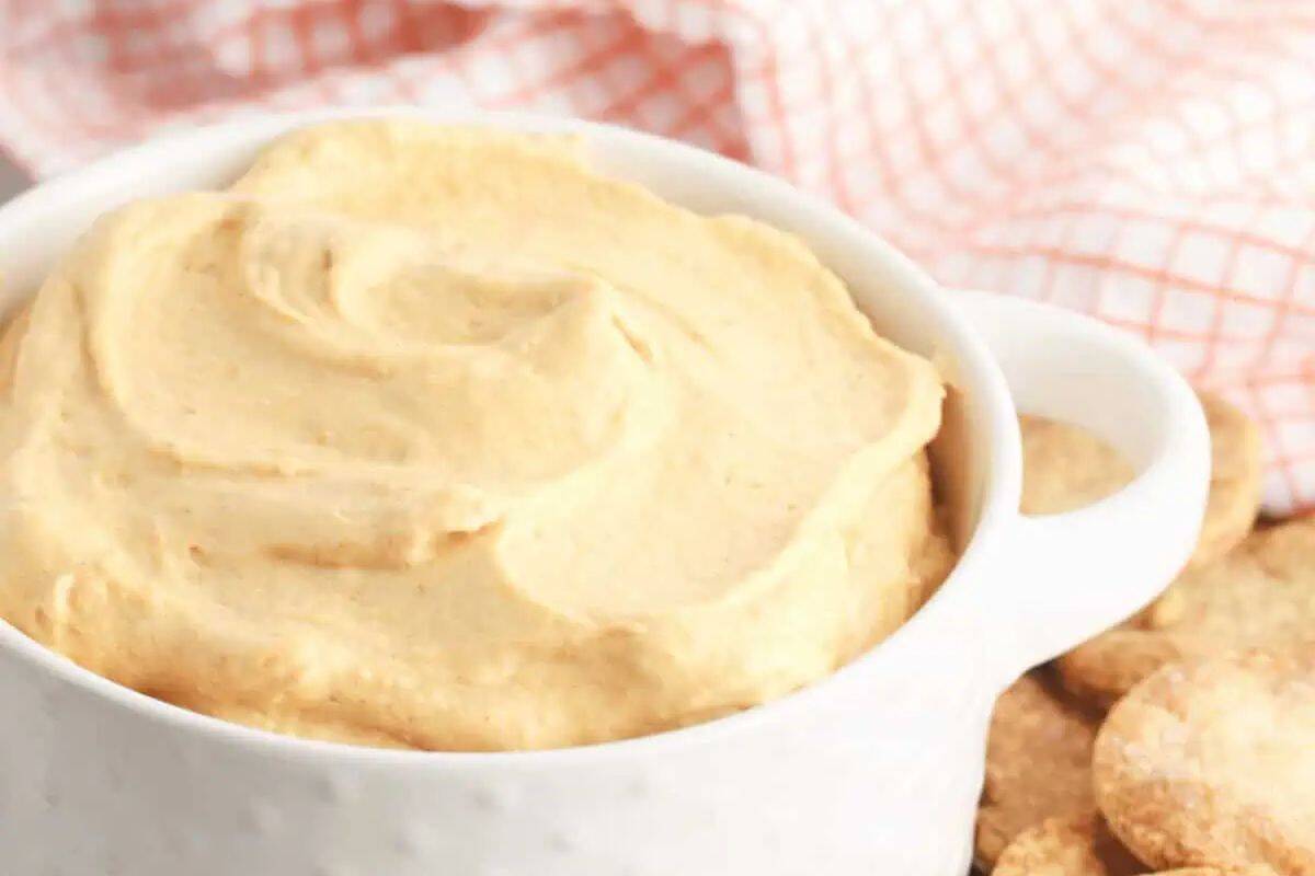 A white bowl filled with creamy, light brown dip sits next to round crackers; a red and white checkered cloth in the background adds cozy fall vibes, perfect for enjoying pumpkin desserts.