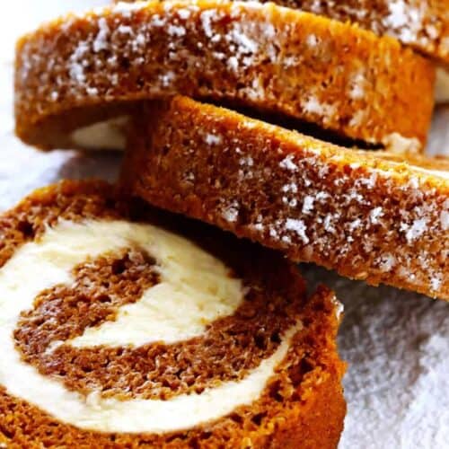 Close-up of several slices of a pumpkin roll cake with cream cheese filling, dusted with powdered sugar, arranged on a white surface—perfect for savoring those cozy Fall Vibes and indulging in delicious Pumpkin Desserts.