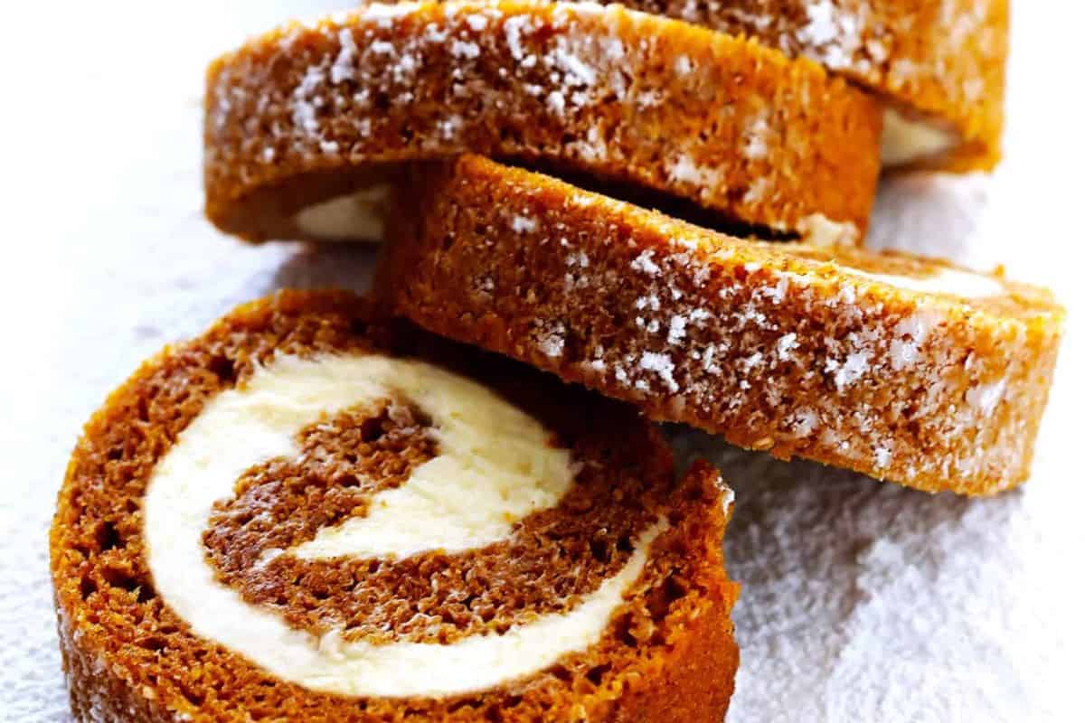 Close-up of several slices of a pumpkin roll cake with cream cheese filling, dusted with powdered sugar, arranged on a white surface—perfect for savoring those cozy Fall Vibes and indulging in delicious Pumpkin Desserts.
