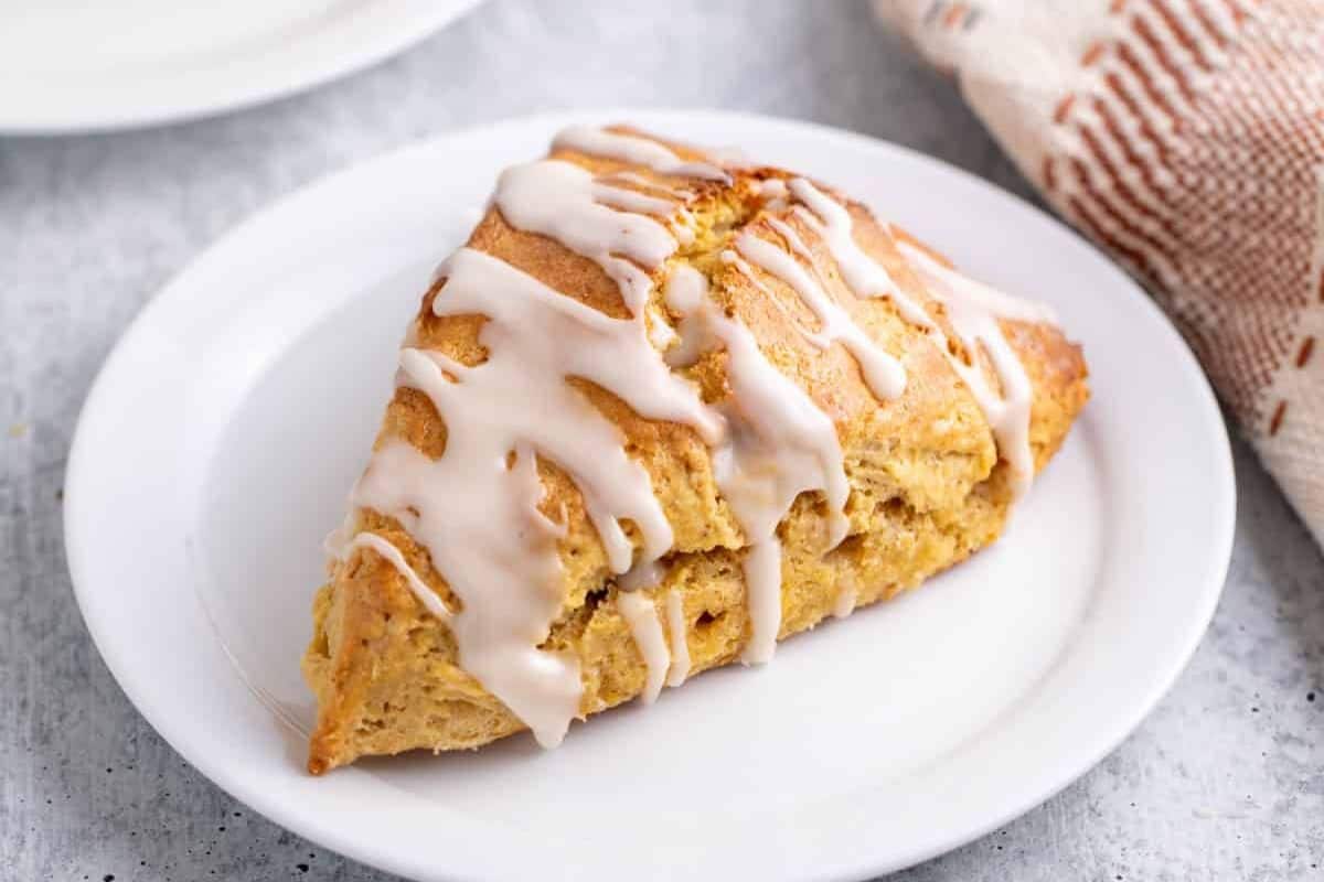 A triangular scone with white icing drizzle sits on a white plate, next to a textured cloth, capturing cozy fall vibes perfect for those who love fall desserts.