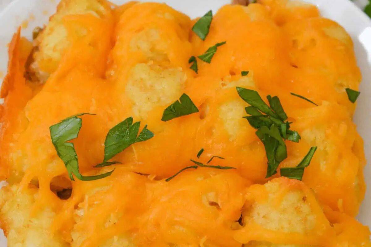Golden tater tots topped with melted cheddar cheese and garnished with chopped parsley on a white plate—perfect for cooler weather or as inspiration for good casserole recipes.