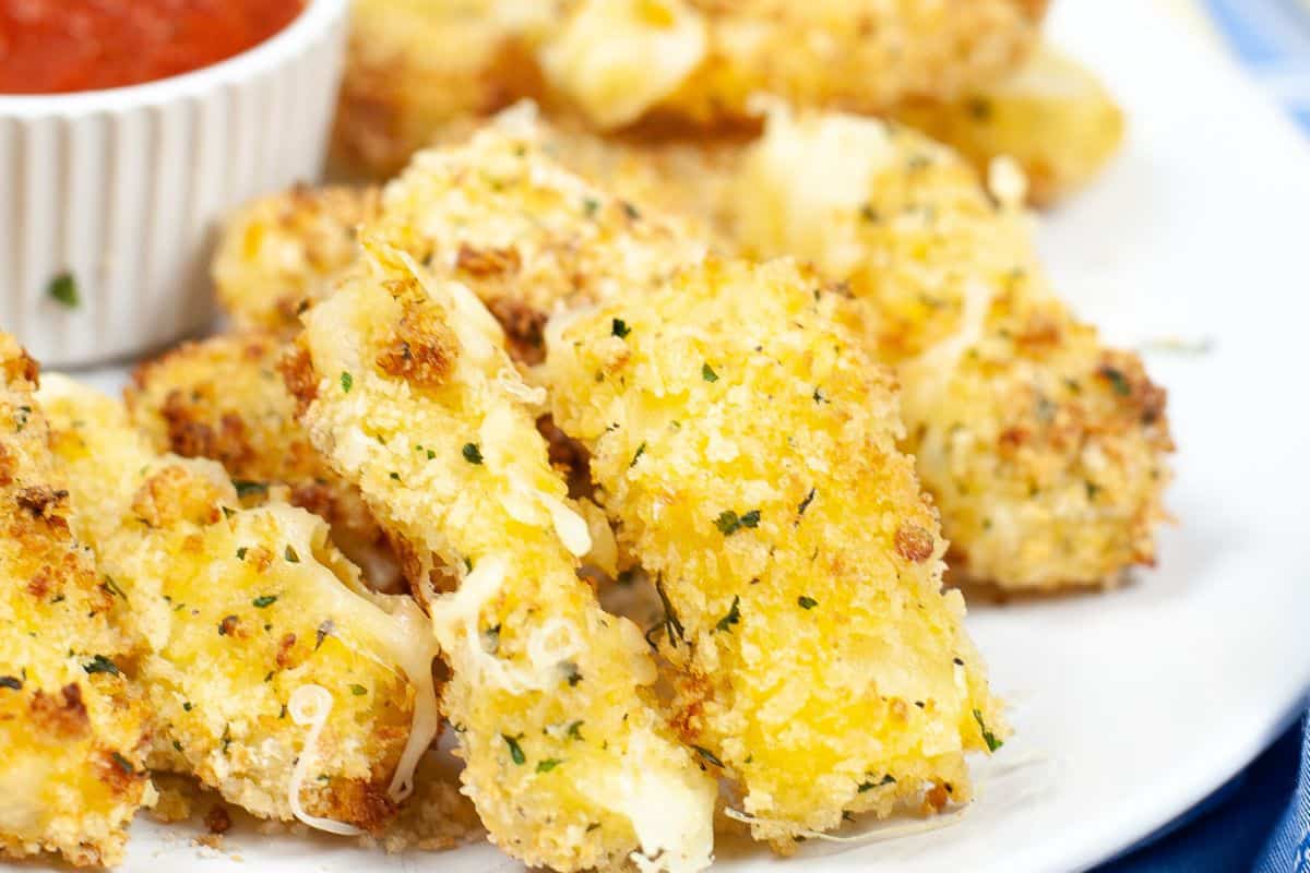 Breaded mozzarella sticks, perfect as a touchdown treat, are piled on a white plate with a small dish of marinara sauce in the background.