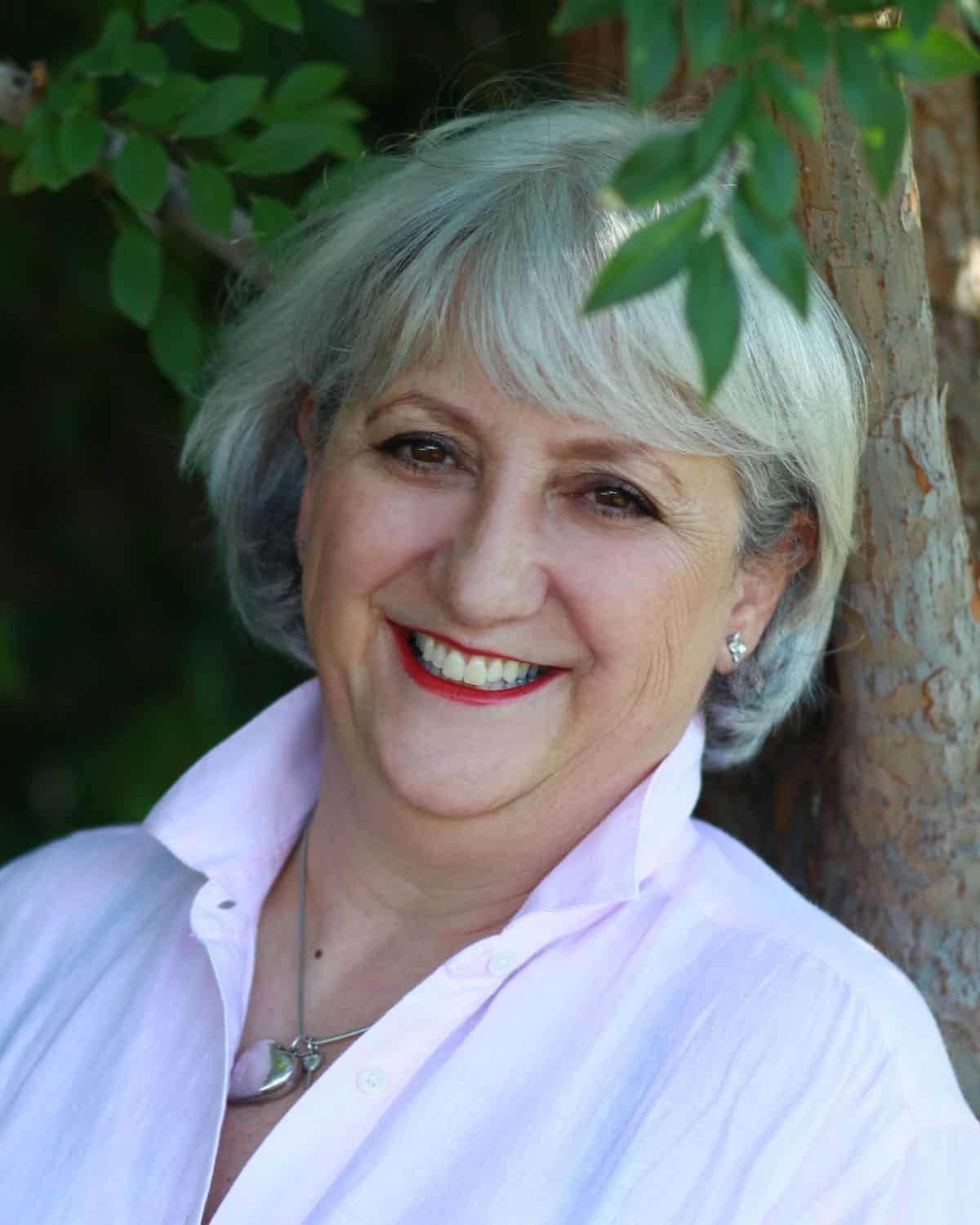A woman with short gray hair and a white shirt smiles warmly while standing outdoors next to a tree, surrounded by greenery. Her joy reflects the contentment she finds in sharing Italian American recipes with loved ones.