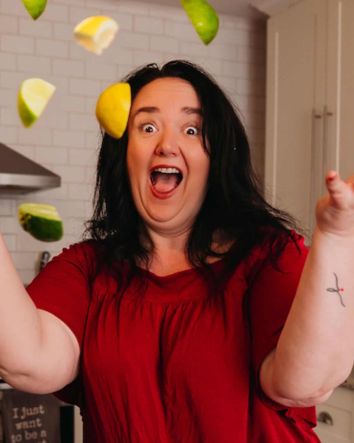 Heather Johnson from the Food Hussy in a red shirt smiles widely while tossing lime and lemon wedges in a kitchen.