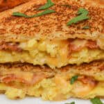 A close-up of a delectable Bacon Egg and Cheese Grilled Cheese sandwich showcases melted cheese, scrambled eggs, and crispy bacon, garnished with parsley on a white plate.