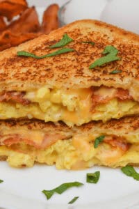 A close-up of a delectable Bacon Egg and Cheese Grilled Cheese sandwich showcases melted cheese, scrambled eggs, and crispy bacon, garnished with parsley on a white plate.