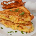 A Bacon, Egg, and Cheese Grilled Cheese sandwich sits enticingly on a white plate, featuring gooey melted cheese between crispy bacon and fluffy scrambled eggs. In the background, cooked bacon and fresh eggs in a glass bowl await their turn to transform into breakfast bliss.