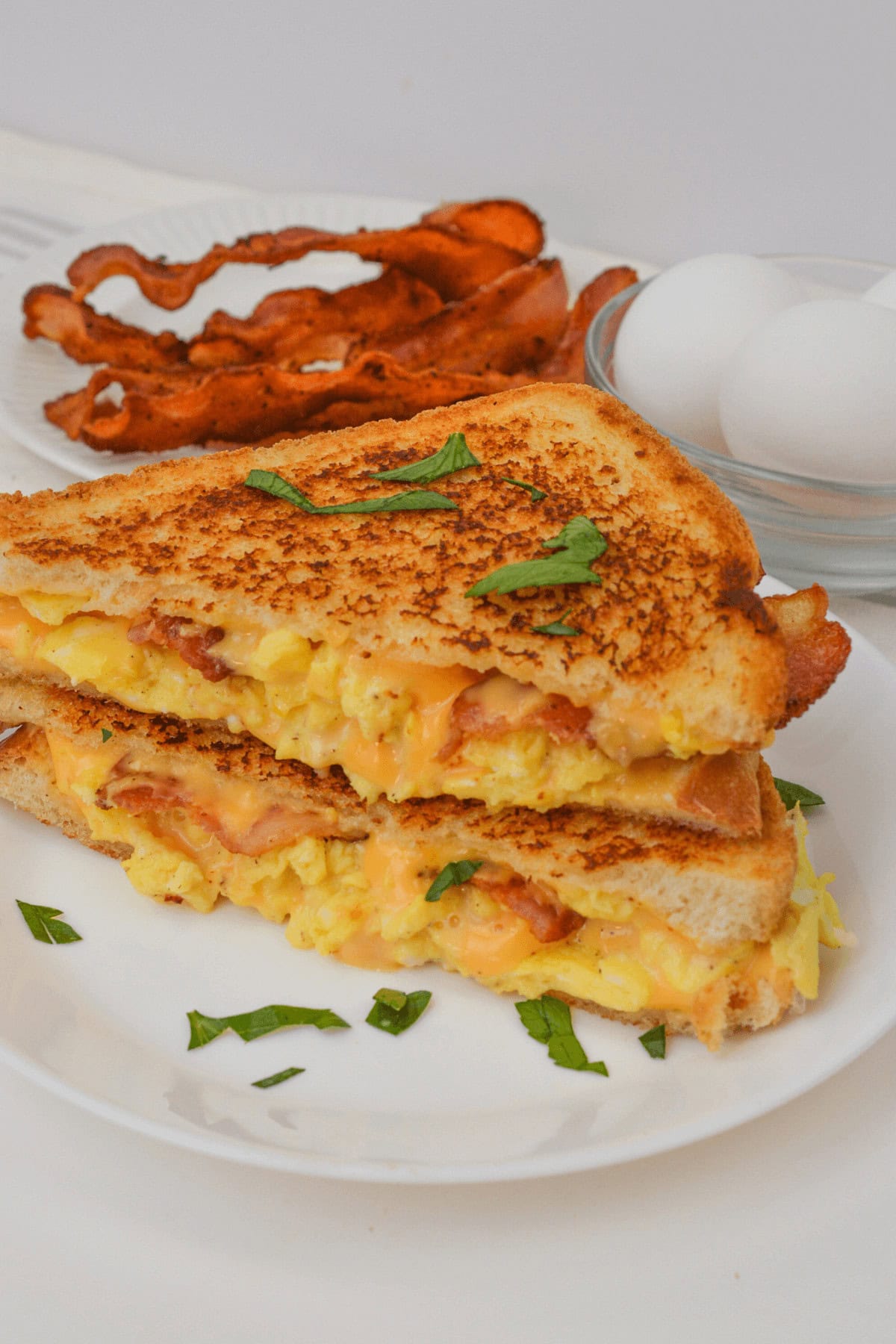 A Bacon, Egg, and Cheese Grilled Cheese sandwich sits enticingly on a white plate, featuring gooey melted cheese between crispy bacon and fluffy scrambled eggs. In the background, cooked bacon and fresh eggs in a glass bowl await their turn to transform into breakfast bliss.
