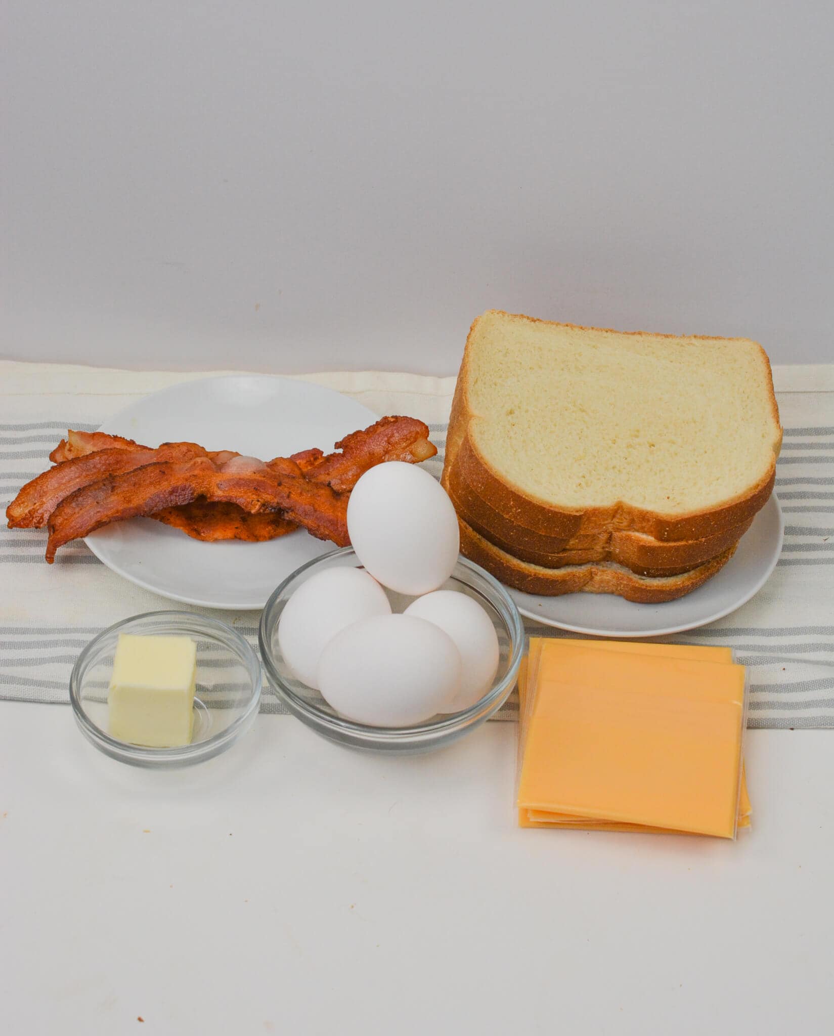 Ingredients: crispy bacon strips, sliced bread, perfectly cooked eggs, rich butter, and melty cheese slices all laid out on the table.