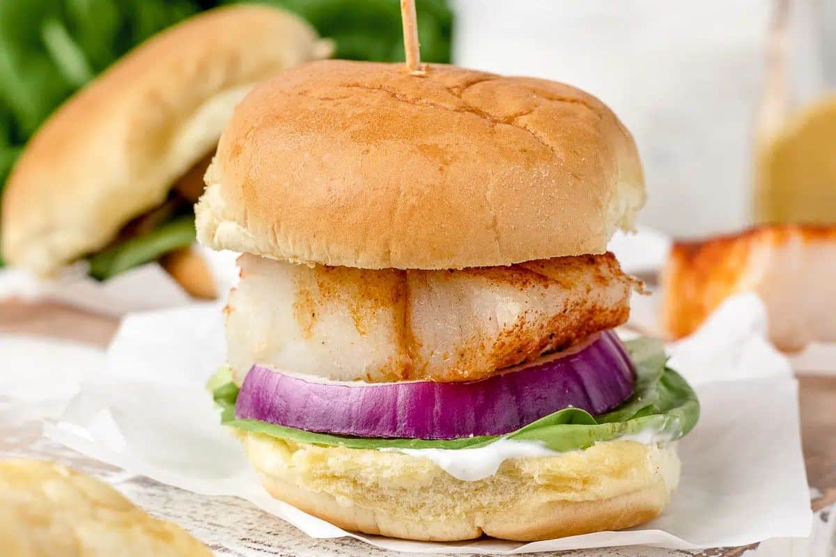 A close-up of a fish slider on a white paper liner, featuring a golden-brown piece of baked cod, a thick slice of red onion, and green lettuce, all on a bun with a creamy white sauce. A wooden toothpick is inserted into the top of the bun.