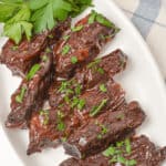 A white plate with glazed, dark-brown country style beef ribs garnished with chopped parsley. A sprig of fresh parsley is placed beside the ribs.