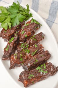 A white plate with glazed, dark-brown country style beef ribs garnished with chopped parsley. A sprig of fresh parsley is placed beside the ribs.