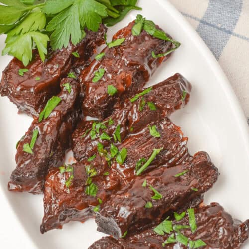 A white plate with glazed, dark-brown country style beef ribs garnished with chopped parsley. A sprig of fresh parsley is placed beside the ribs.