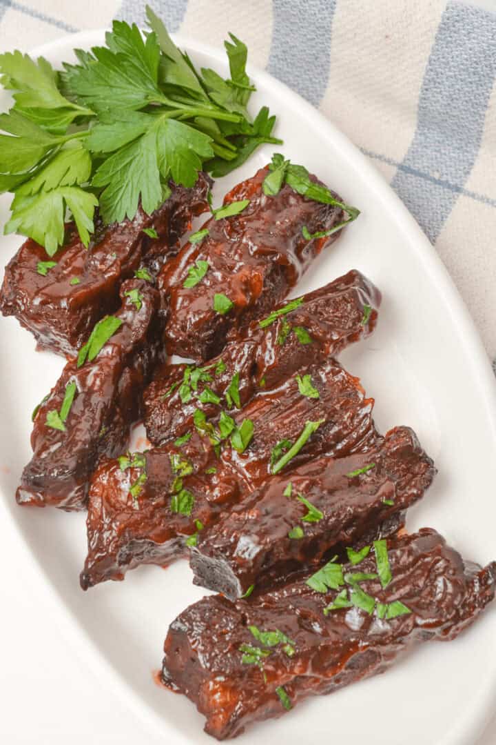 A white plate with glazed, dark-brown country style beef ribs garnished with chopped parsley. A sprig of fresh parsley is placed beside the ribs.
