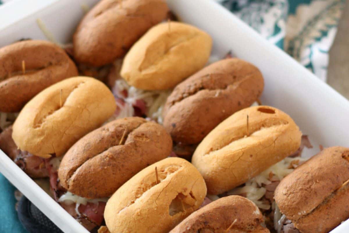 An overhead, slightly angled shot of a white baking dish filled with multiple small, oval-shaped baked pastrami sandwiches with toothpicks inserted into their tops. The buns are a mix of light and dark brown, and shredded cheese is visible underneath.