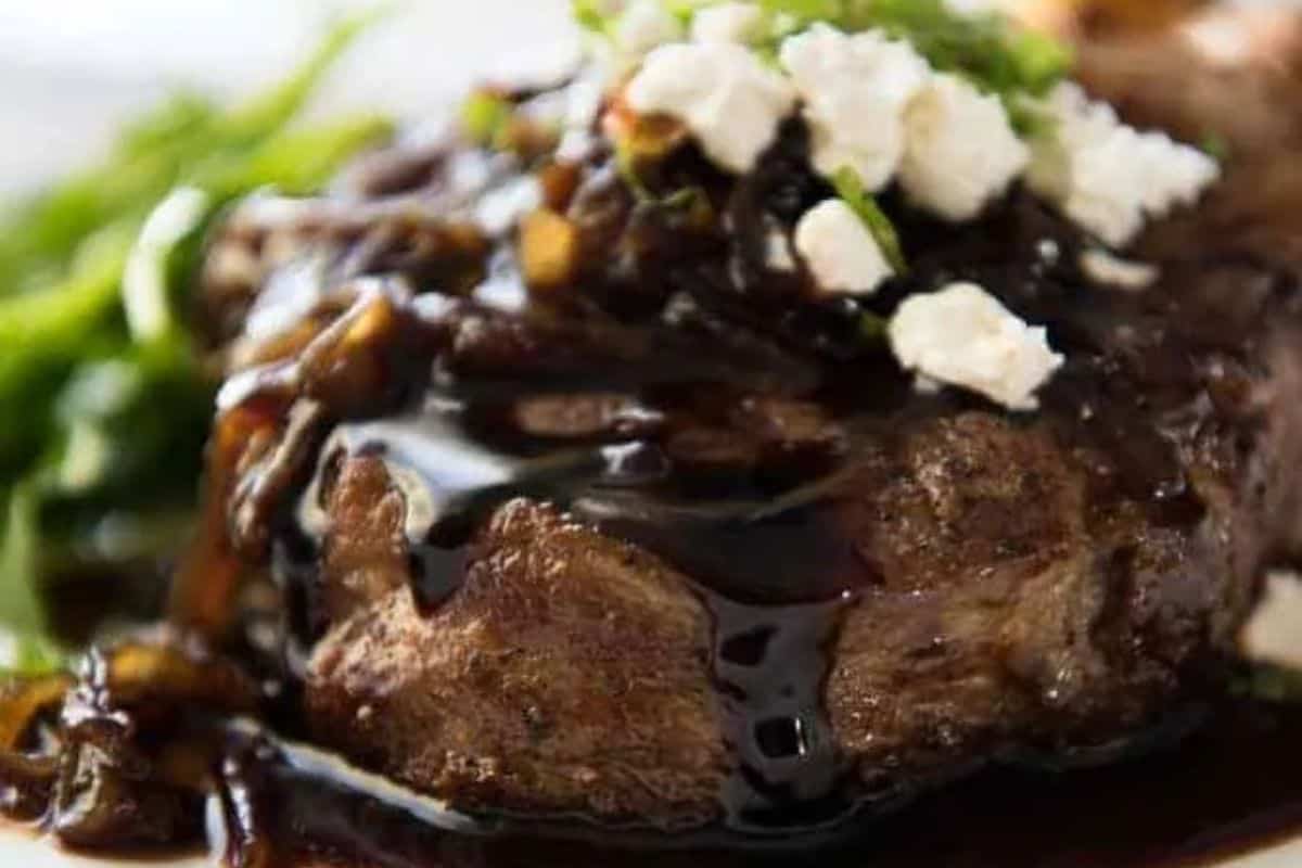 Seared pork chop topped with caramelized onions and drizzled with a rich balsamic glaze, garnished with crumbled feta and fresh herbs.