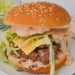 A Big Mac-inspired burger with a sesame seed bun, crisp lettuce, melted cheese, ground beef, and tangy pickles, all drizzled with special sauce and served on a white plate.