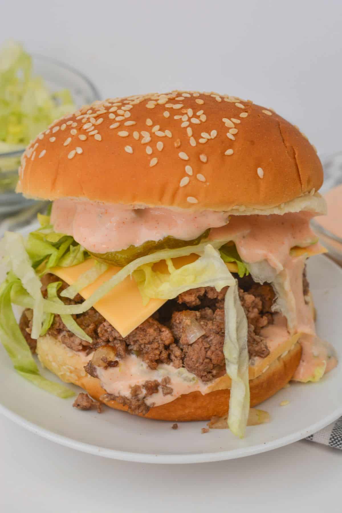 A Big Mac-inspired burger with a sesame seed bun, crisp lettuce, melted cheese, ground beef, and tangy pickles, all drizzled with special sauce and served on a white plate.