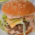 Close-up of a sesame seed hamburger bun with layers of lettuce, cheese, pickle slices, and savory ground beef smothered in sauce on a plate. It’s like a gourmet twist on Big Mac flavors. A bowl of fresh lettuce lurks in the background for the perfect Sloppy Joes companion.