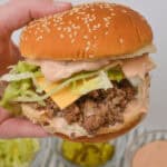 A hand holds a Big Mac sloppy joes, with lettuce, cheese, ground beef, and sauce cascading down the sides. Bowls of toppings are visible in the background, offering endless possibilities to create your own version of Sloppy Joes.