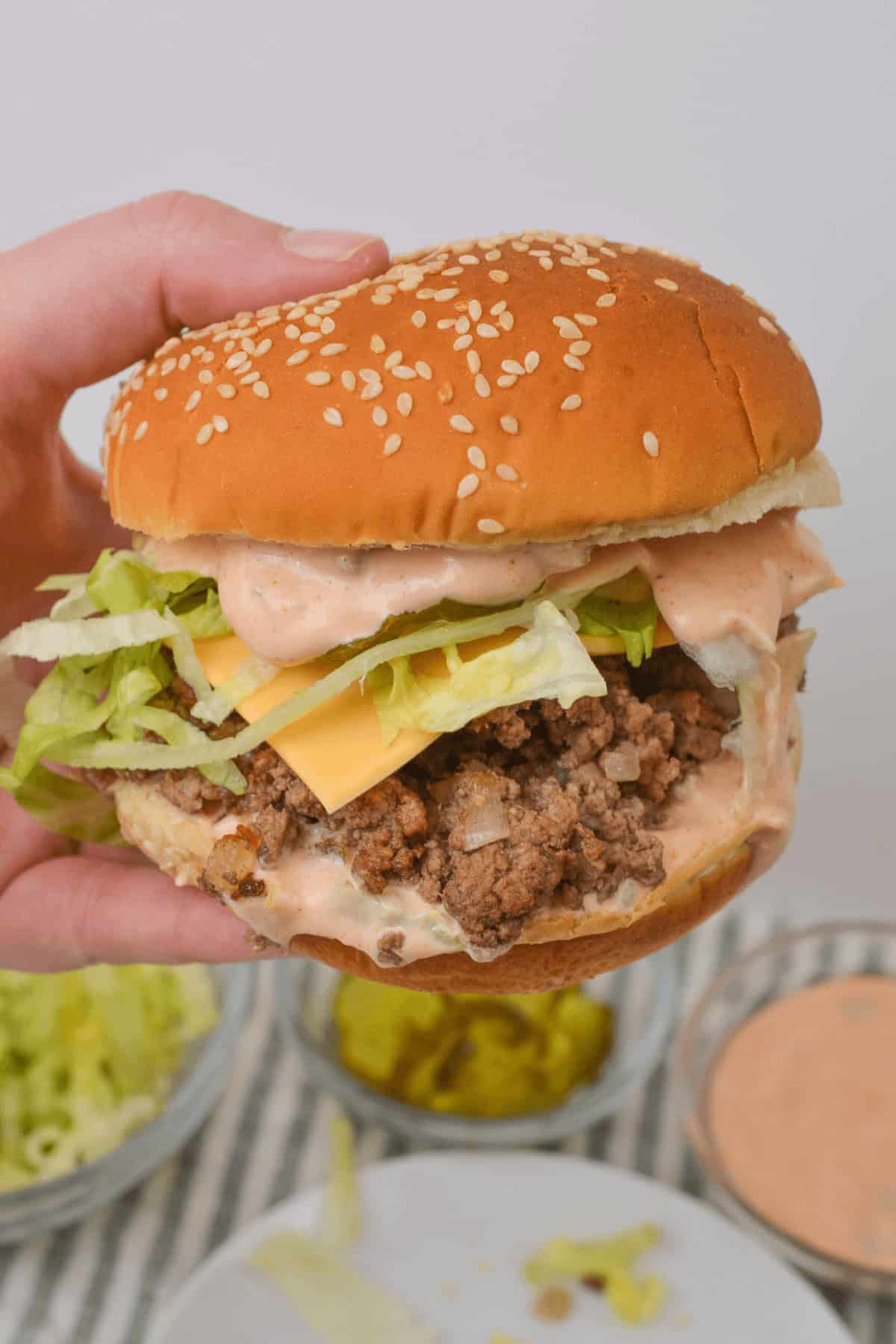 A hand holds a Big Mac sloppy joes, with lettuce, cheese, ground beef, and sauce cascading down the sides. Bowls of toppings are visible in the background, offering endless possibilities to create your own version of Sloppy Joes.
