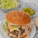 A sesame seed bun burger resembling a Big Mac, complete with lettuce, cheese, and sauce on a plate. Nearby are bowls of shredded lettuce, pickles, and extra sauce on a striped cloth.