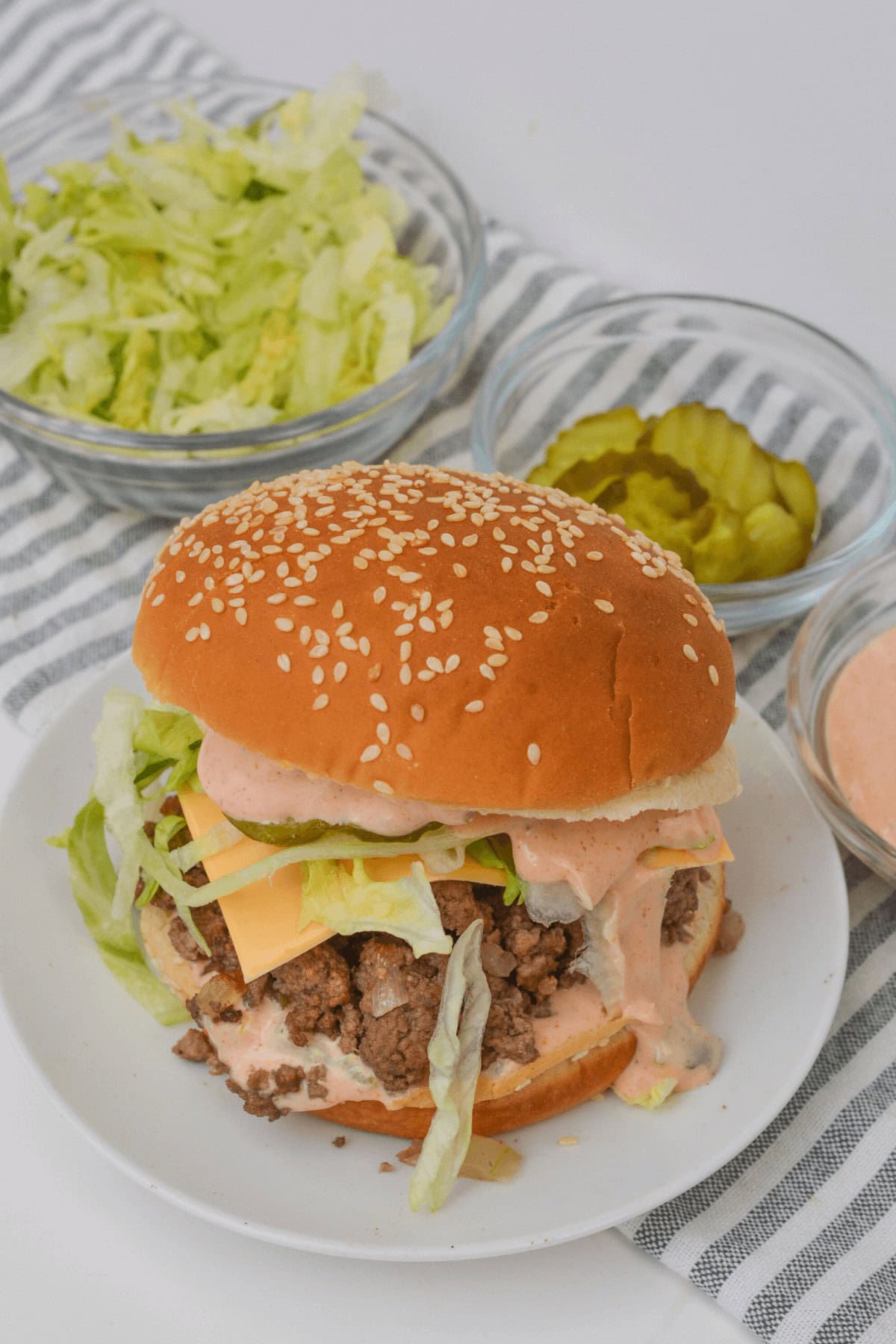 A sesame seed bun burger resembling a Big Mac, complete with lettuce, cheese, and sauce on a plate. Nearby are bowls of shredded lettuce, pickles, and extra sauce on a striped cloth.