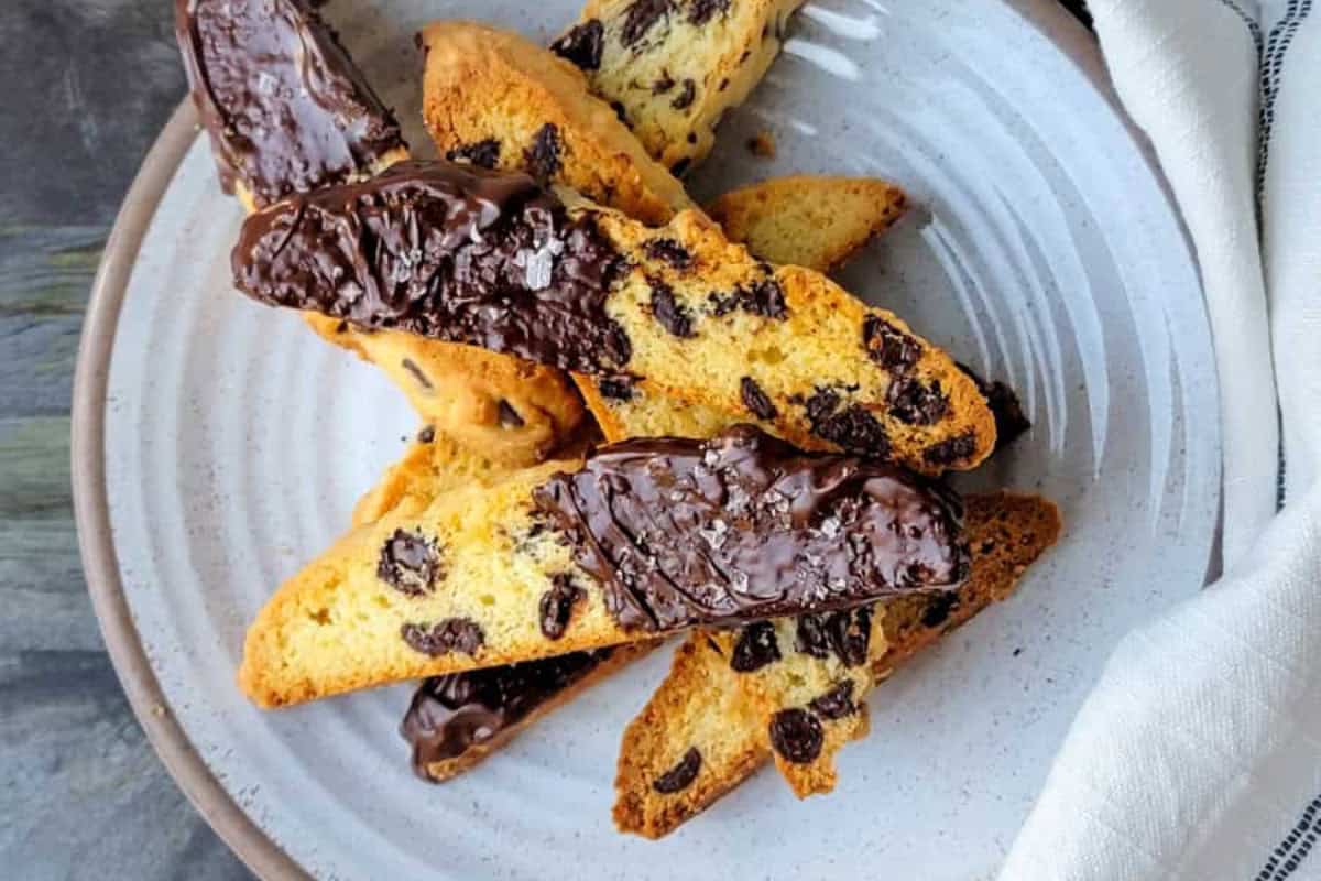 A plate of chocolate-dipped biscotti with visible chocolate chips, arranged in a stack on a white ceramic plate next to a folded napkin—perfect for adding to your favorite chocolate recipes or an indulgent desserts collection.