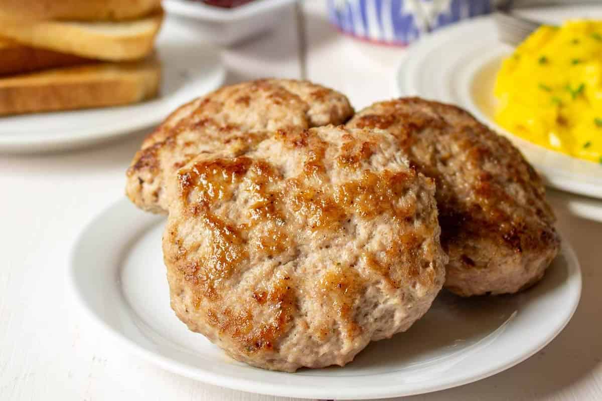 Three cooked sausage patties on a white plate with toast and scrambled eggs in the background—perfect for weekend brunch or adding to your favorite lazy morning recipes.