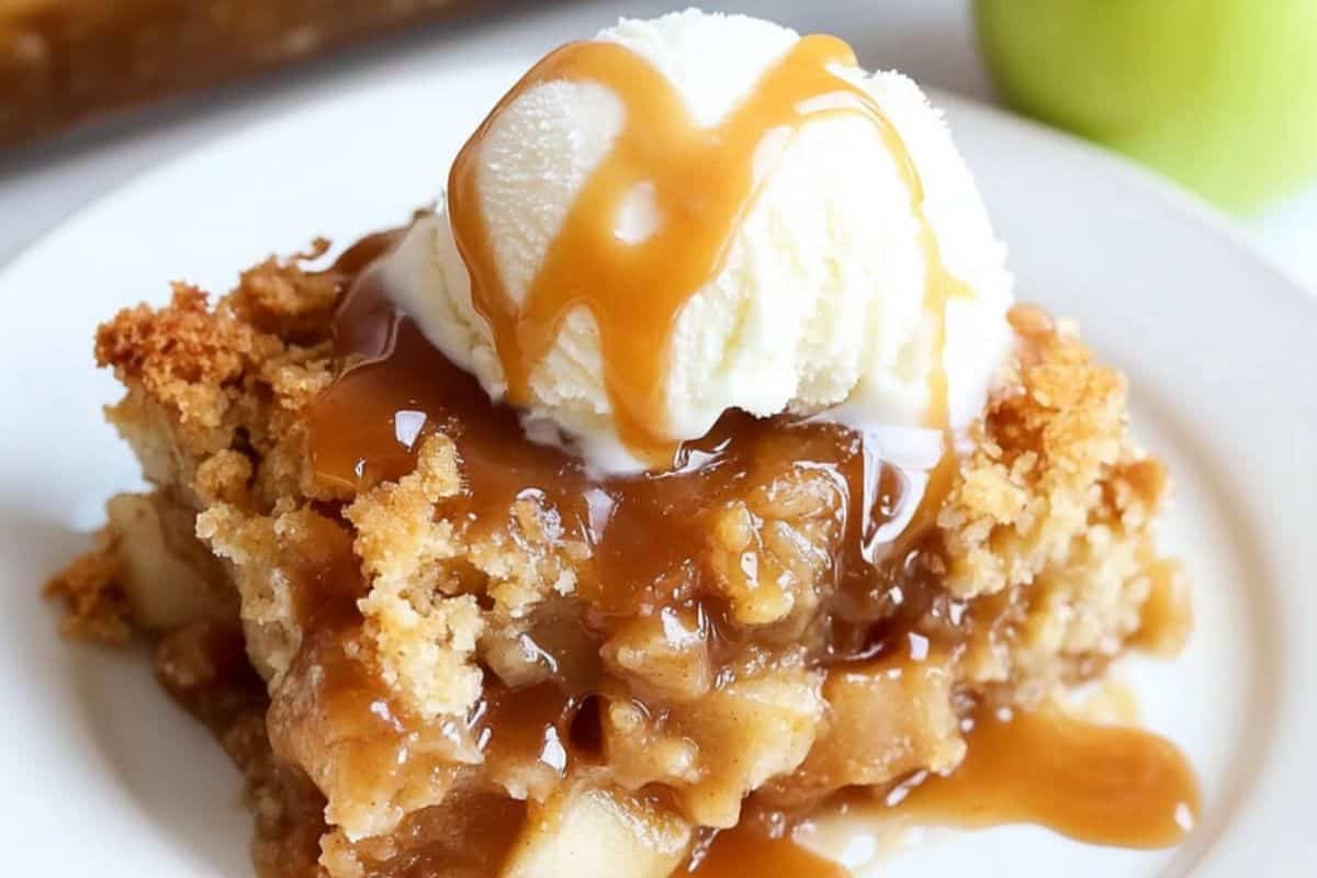 Close-up of a slice of caramel apple dump cake, served on a white plate and topped with vanilla ice cream and caramel drizzle.