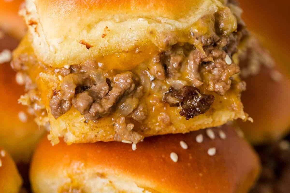 A close-up of a cheeseburger slider, stacked on top of another slider, showing a generous amount of ground meat and melted cheese, with sesame seeds visible on the buns.
