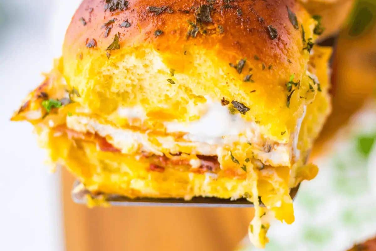  A close-up of a golden-brown slider topped with green herbs, being lifted by a metal spatula, revealing layers of chicken, bacon, and creamy ranch sauce with melted cheese dripping down.