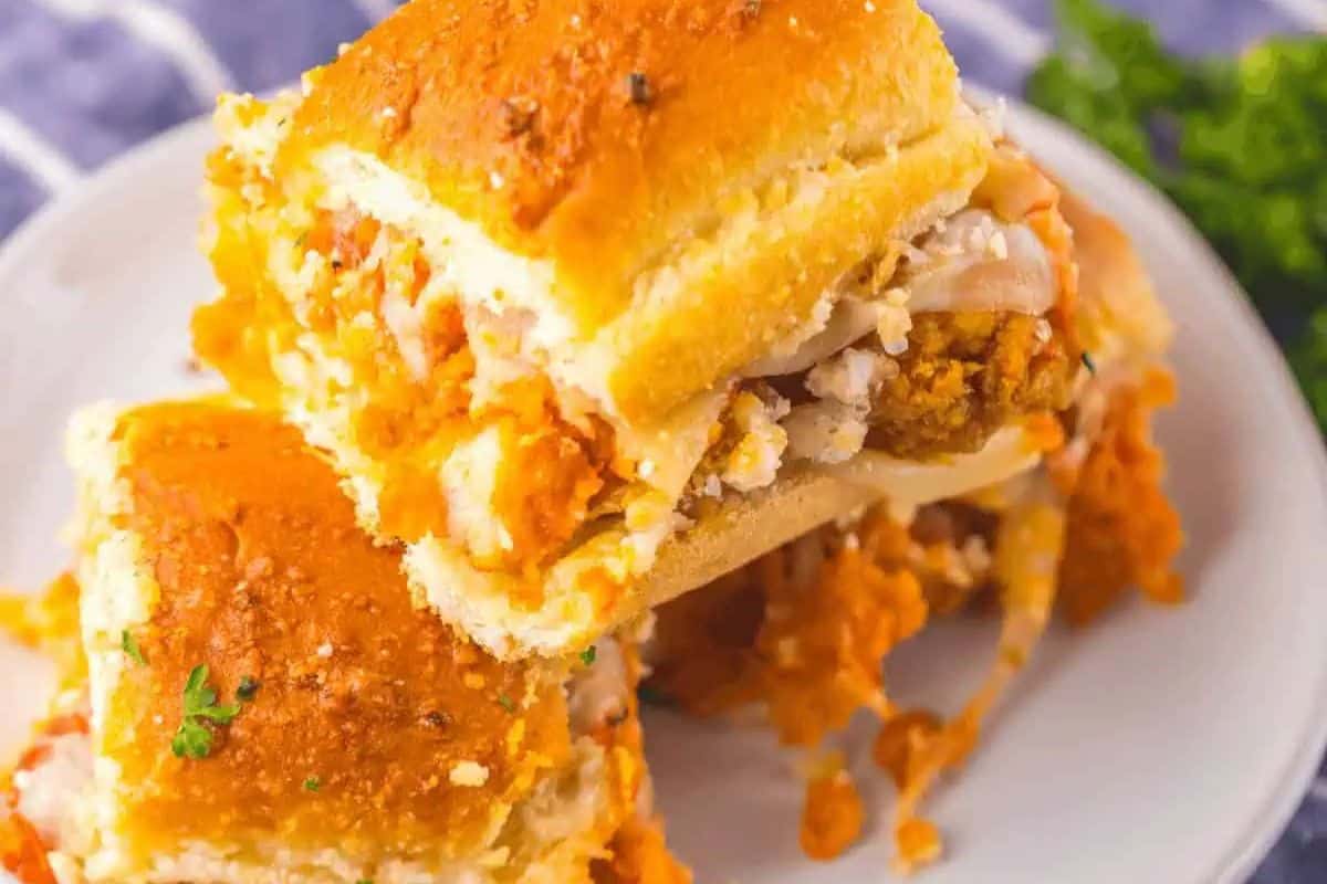 A close-up of several chicken parmesan sliders stacked on a white plate, showing golden-brown chicken pieces, melted white cheese, and a red sauce, all on soft buns.