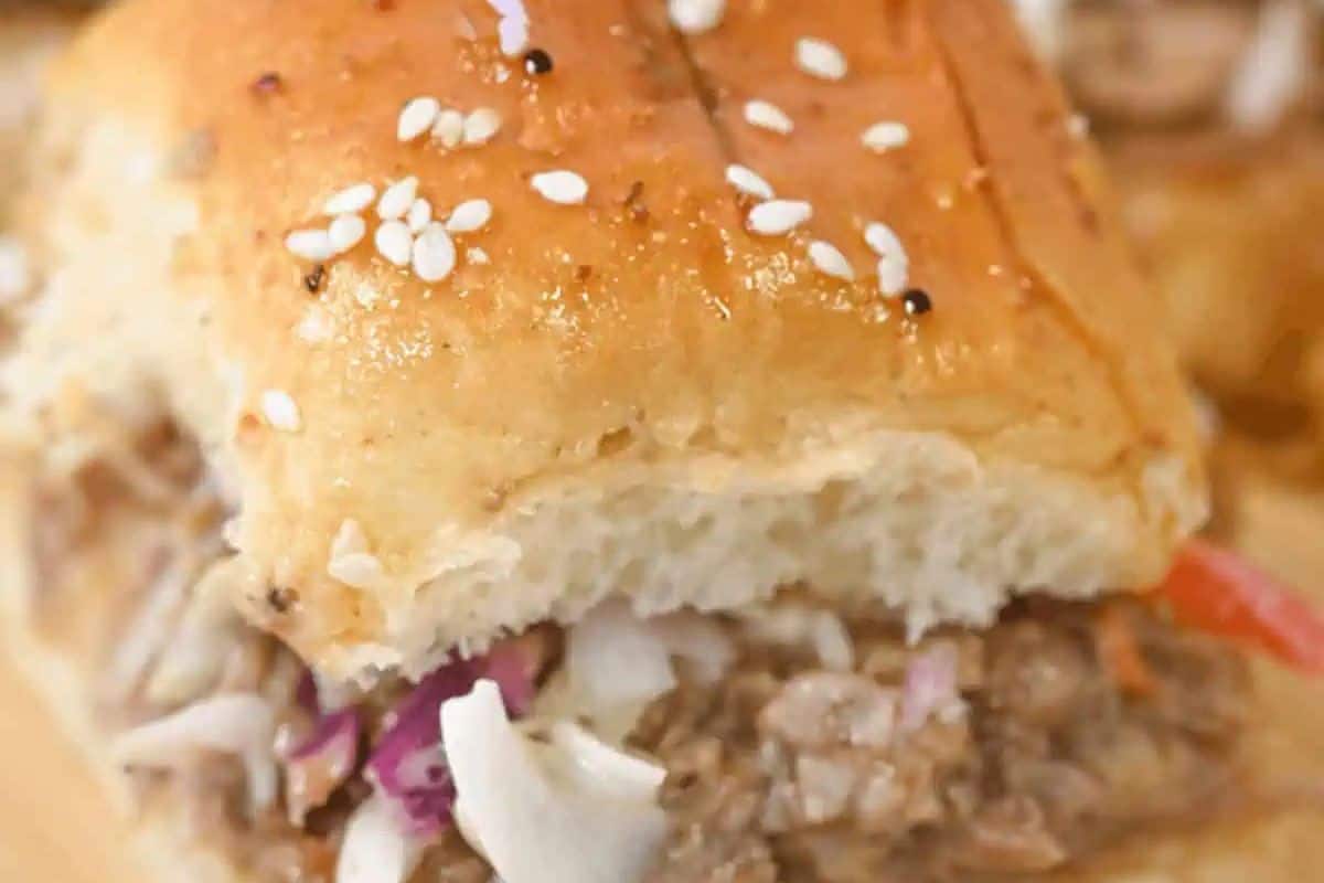  A close-up of a slider bun topped with white sesame seeds, revealing a filling of shredded pork and a colorful coleslaw with red cabbage.