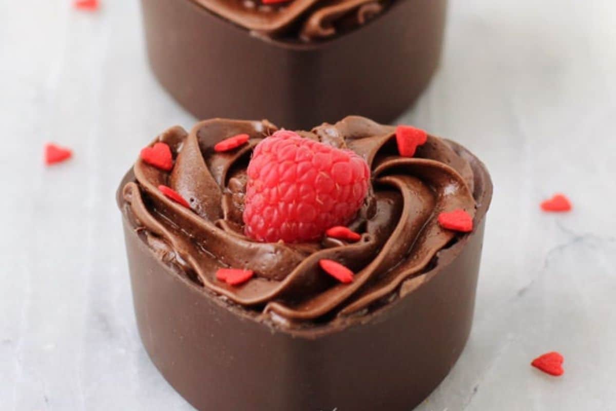 A Valentine sweetheart delight: heart-shaped chocolate dessert with swirled frosting, topped by a raspberry and tiny red heart sprinkles, resting on a light surface. Perfect for your Valentine dessert ideas.