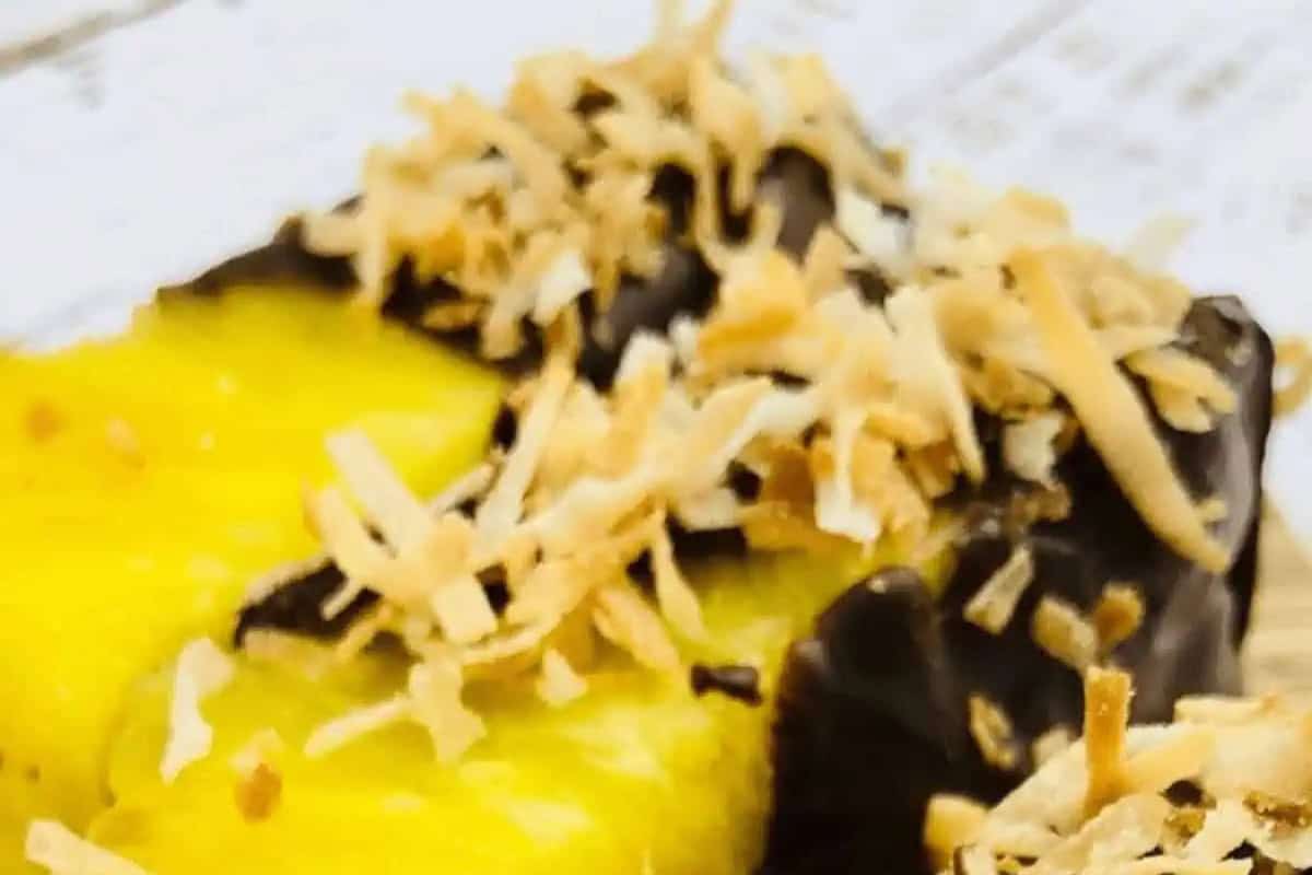 A slice of pineapple is partially dipped in chocolate and topped with toasted shredded coconut on a light-colored surface—an irresistible treat for any chocolate recipes collection.
