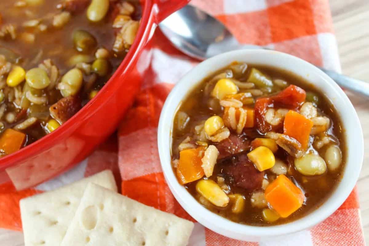 Cozy up and warm up with a bowl of vegetable soup filled with corn, carrots, and beans, served next to saltine crackers and a spoon on an orange plaid cloth.