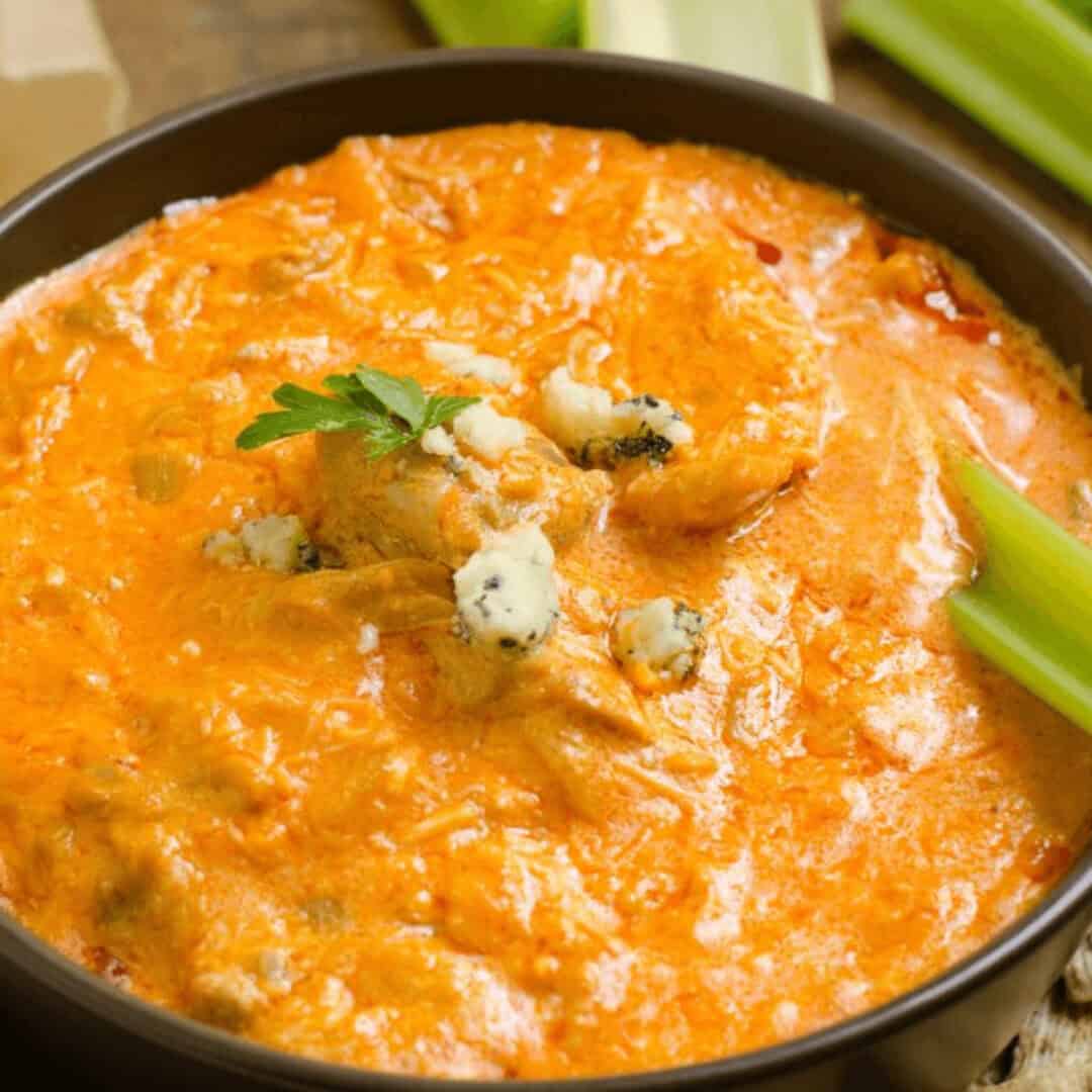 A bowl of creamy Cottage Cheese Buffalo Chicken Dip topped with blue cheese crumbles, garnished with parsley, served with crisp celery sticks on the side.