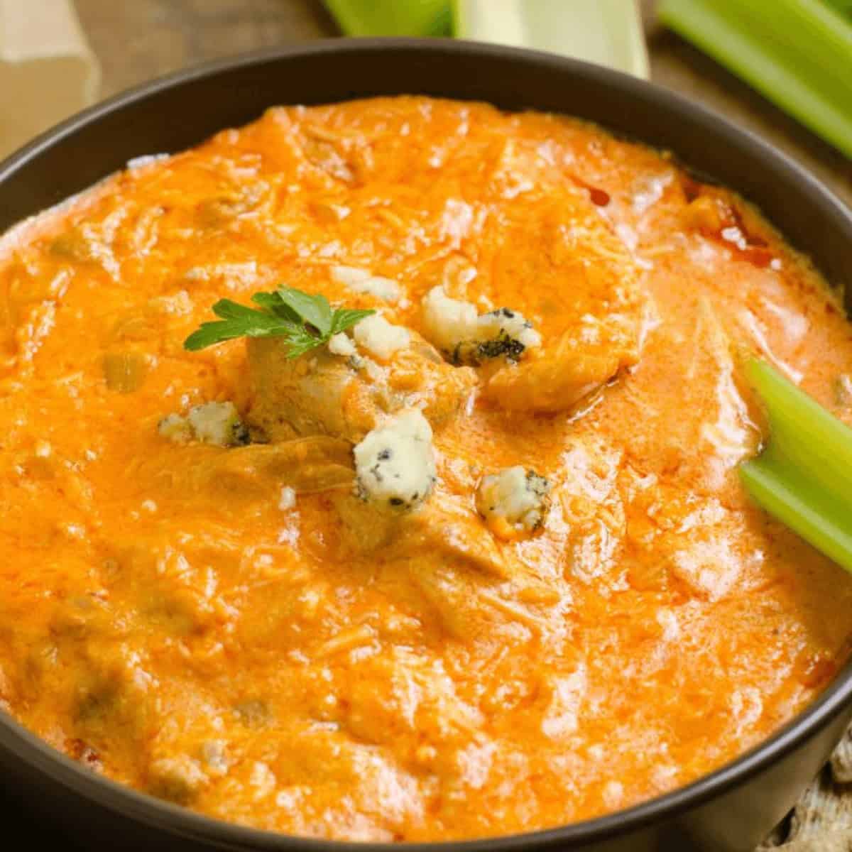 A bowl of creamy Cottage Cheese Buffalo Chicken Dip topped with blue cheese crumbles, garnished with parsley, served with crisp celery sticks on the side.