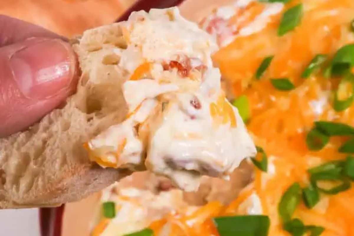 A hand holding a piece of bread with a creamy cheese dip, featuring diced vegetables and garnished with green onions in the background. This irresistible dip is perfect for scoring big points at your Super Bowl party!.