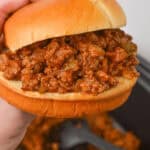 A hand holding a Crock Pot Sloppy Joe sandwich filled with a seasoned ground meat mixture, with more filling simmering in the background.