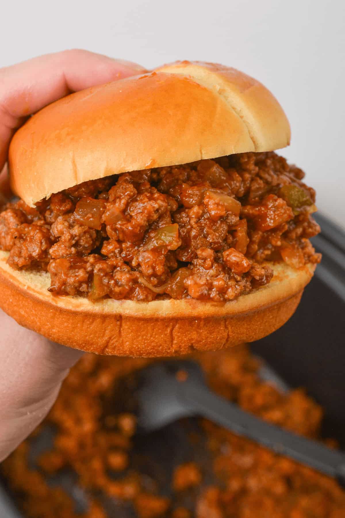 A hand holding a Crock Pot Sloppy Joe sandwich filled with a seasoned ground meat mixture, with more filling simmering in the background.