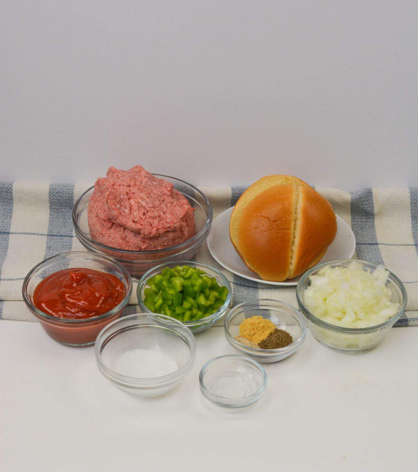 Ground meat, a hamburger bun, chopped onion, green bell pepper, ketchup, and an array of spices sit in bowls on a cloth, ready to transform into delicious recipe.
