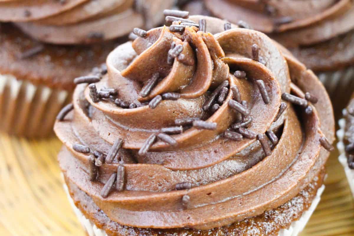 Close-up of a chocolate cupcake topped with swirled chocolate frosting and chocolate sprinkles, part of another cupcake visible in the background—a perfect addition to any chocolate collection.