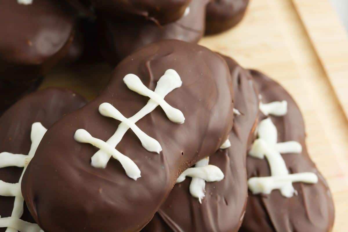 Football-themed recipes come to life with chocolate-covered cookies shaped like footballs, featuring white icing stitches. Perfectly arranged on a wooden surface, these touchdown treats are an ideal addition to any game day party spread.