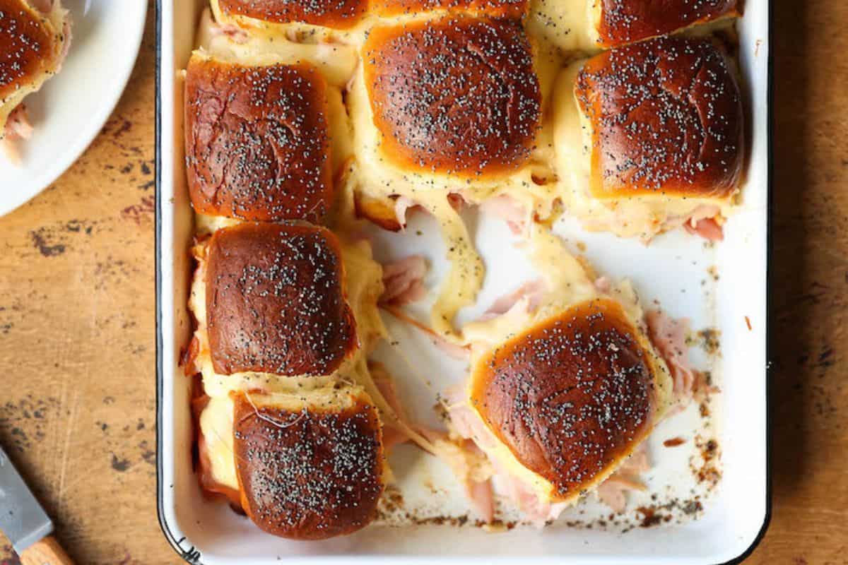  An overhead shot of a baking dish filled with baked ham and swiss sliders, featuring golden-brown buns heavily sprinkled with poppy seeds and melted, gooey cheese spilling out from between the buns.