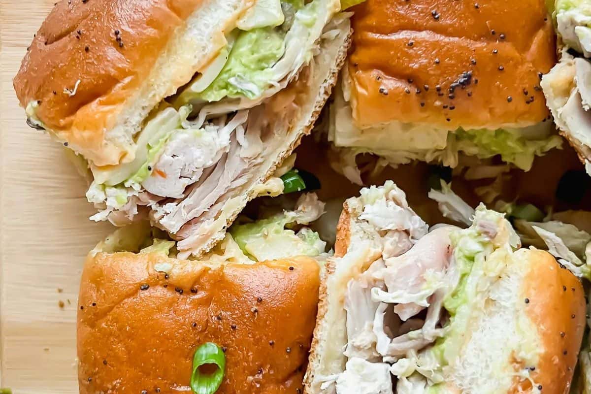 An overhead view of several Hawaiian chicken sliders, some cut in half to show shredded chicken and green slaw on soft buns, with some visible sesame seeds on top.