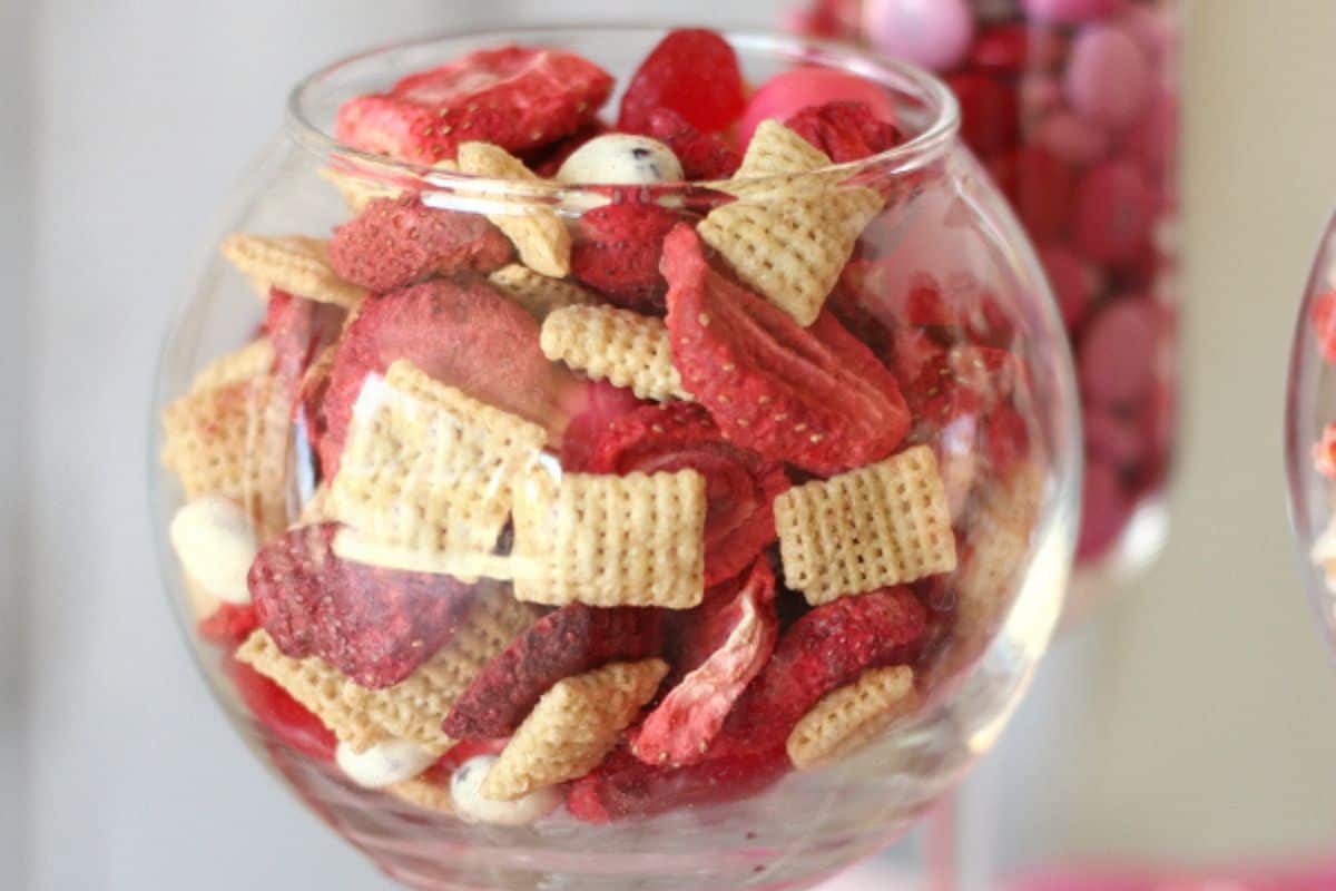 A glass bowl containing cereal squares mixed with dried strawberries and assorted red and pink candies, perfect for Creative Valentine celebrations, offers a delightful treat.