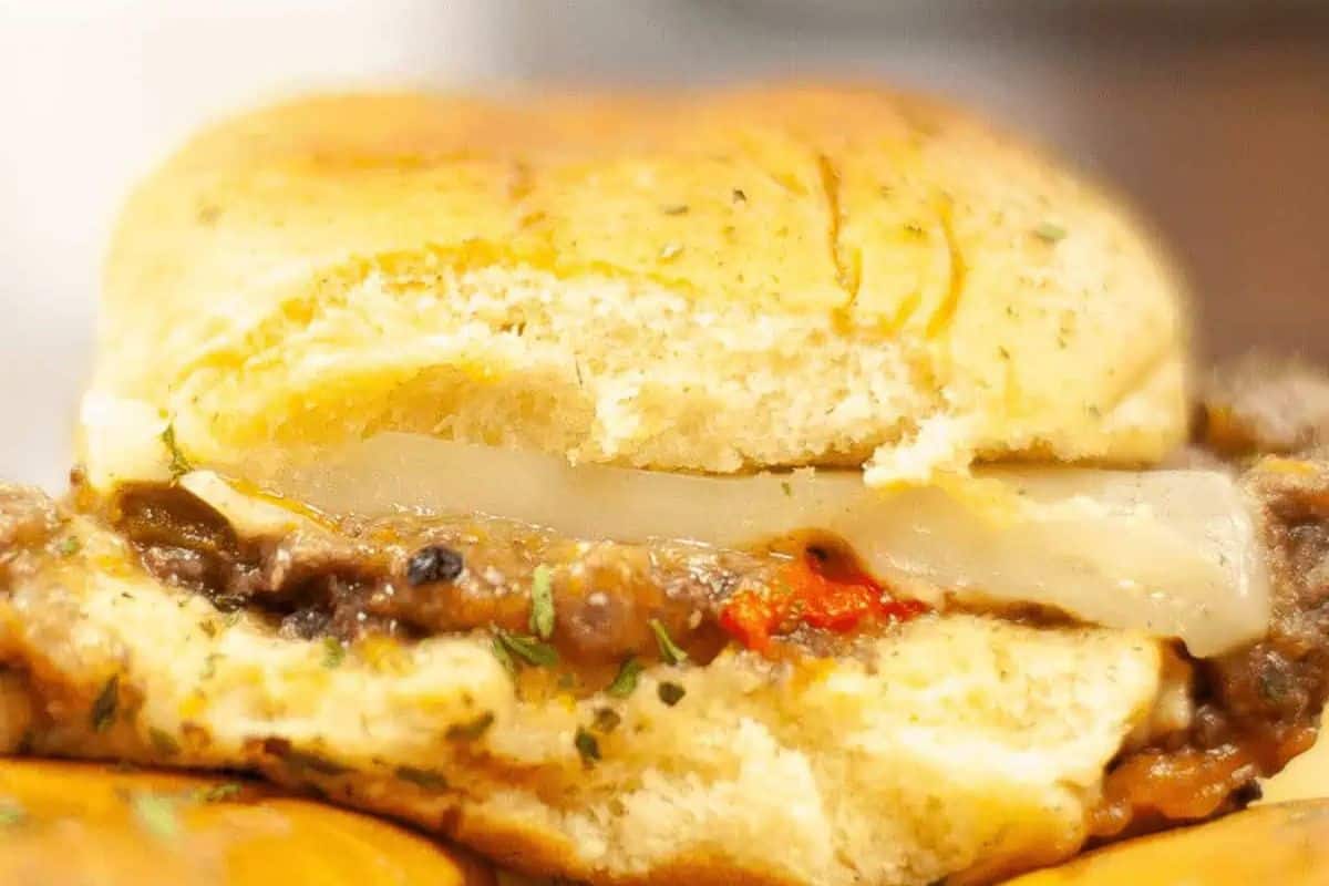 A close-up of a slider with steak and melted white cheese, along with some red and green vegetables, on a golden-brown bun.