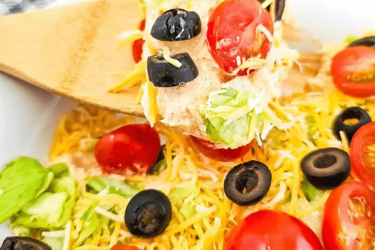 Close-up of a wooden spoon lifting a mix of shredded cheese, cherry tomatoes, olives, and lettuce from a bowl—score big points with this colorful combo that's sure to be a hit at your next Super Bowl party.